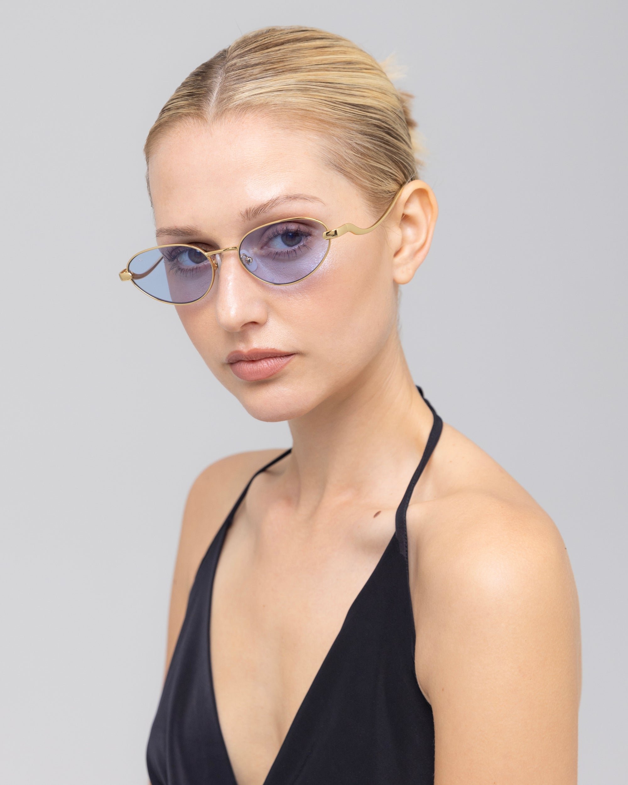A woman with blond hair tied back wears Rebirth blue-tinted oval sunglasses by For Art's Sake®, paired with a black halter top, looking at the camera against a plain light gray background.