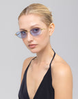 A woman with blond hair tied back wears Rebirth blue-tinted oval sunglasses by For Art's Sake®, paired with a black halter top, looking at the camera against a plain light gray background.