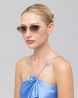 A woman with light skin and blonde hair in a sleek bun models the Rebirth rimless cat-eye sunglasses by For Art's Sake®, wearing a shiny blue spaghetti strap top against a plain light gray background.