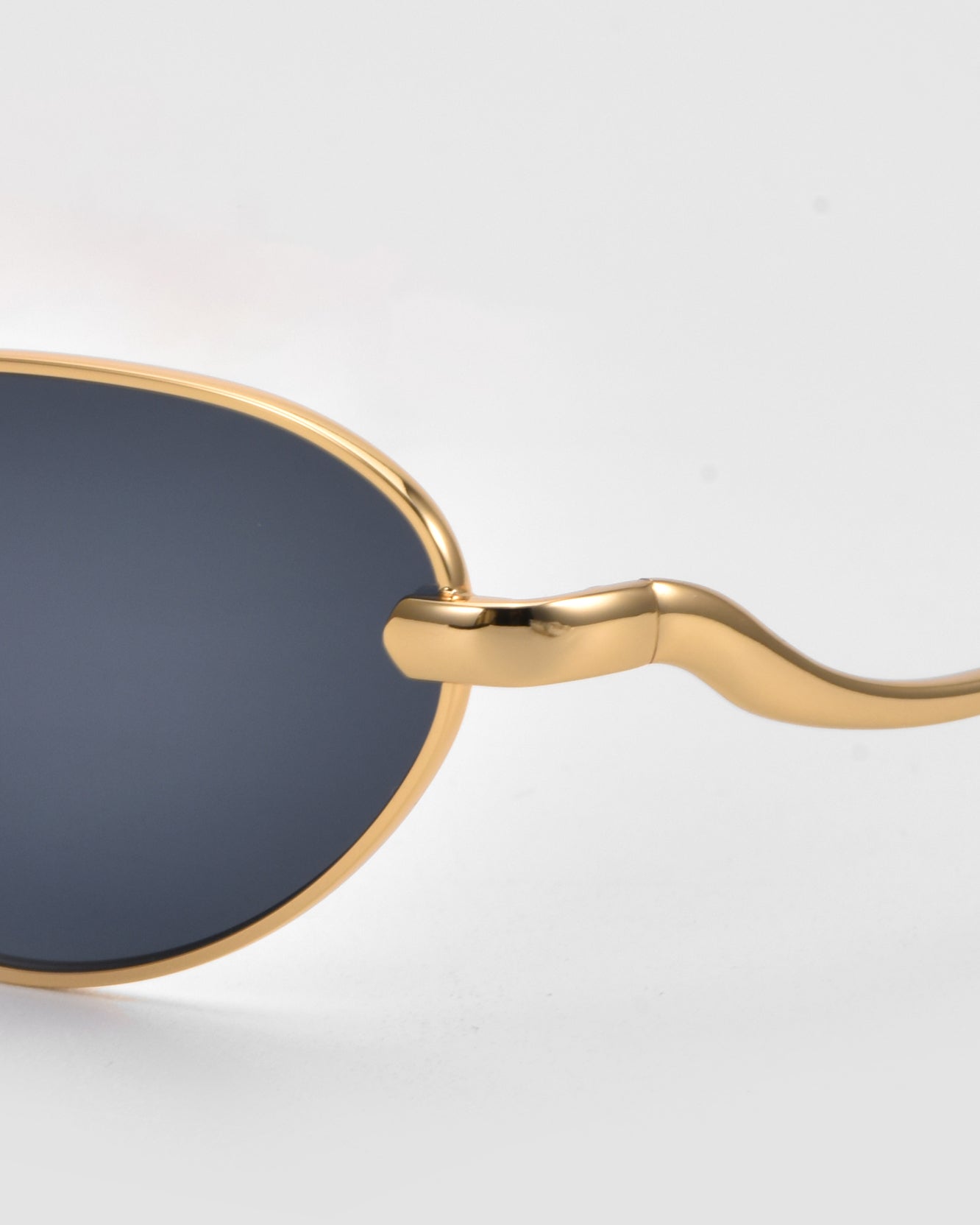 Close-up of Rebirth by For Art&#39;s Sake®: slim oval sunglasses with a gold metal frame, dark-tinted lenses, and uniquely curved temples set against a light gray background—a timeless look for modern style.