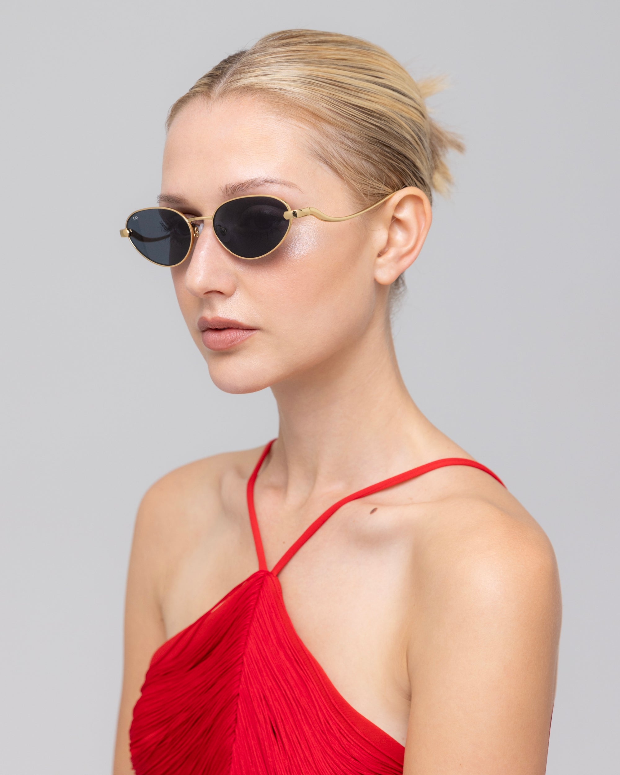 A woman with blonde hair in a bun models For Art's Sake® "Rebirth" futuristic sunglasses and a red halter-neck dress, posing against a plain light gray background while looking slightly to the side.