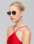 A woman with blonde hair in a bun models For Art's Sake® "Rebirth" futuristic sunglasses and a red halter-neck dress, posing against a plain light gray background while looking slightly to the side.