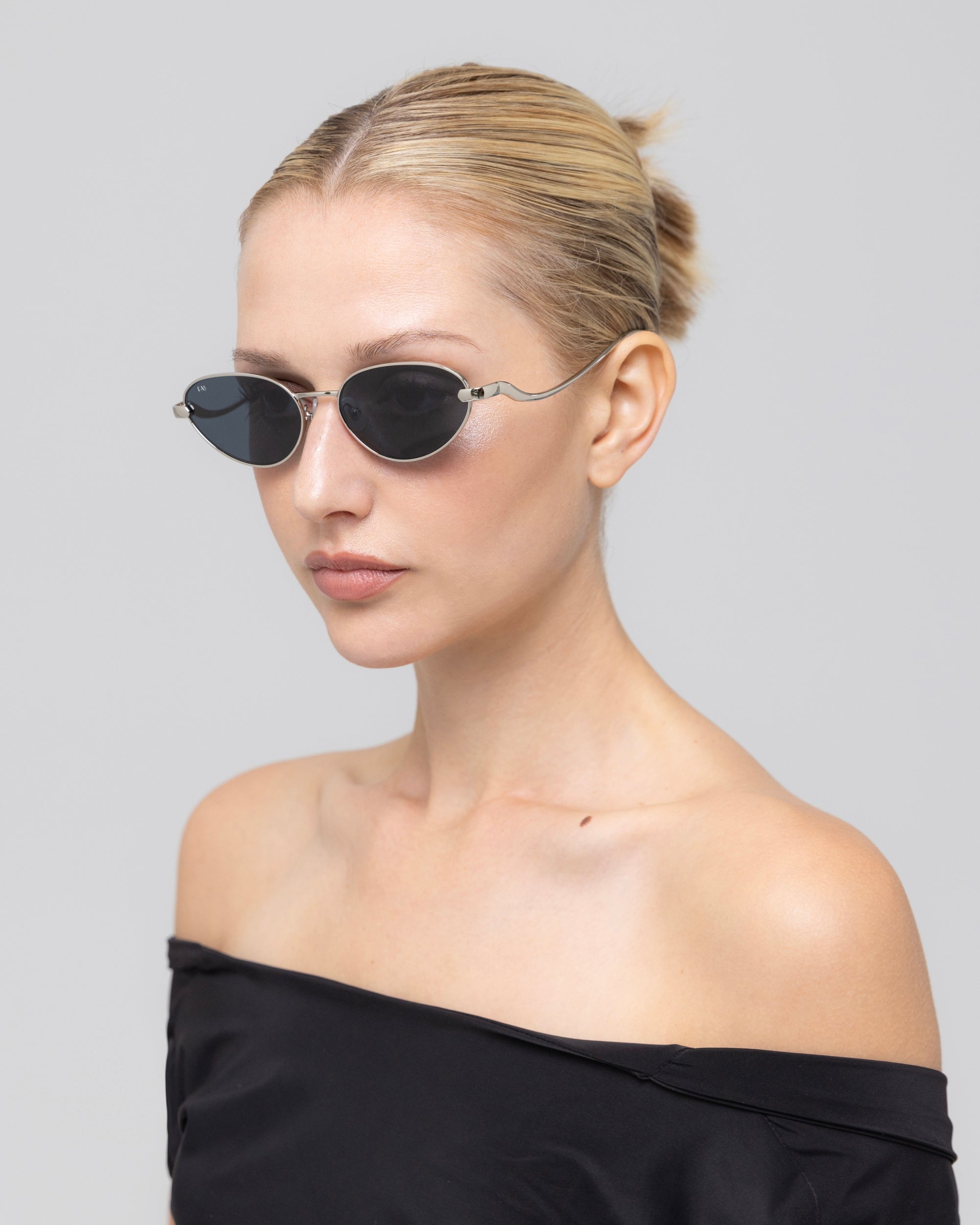 A woman with blonde hair in a bun wears Rebirth sunglasses by For Art's Sake®, paired with an off-the-shoulder black top, posing against a plain light gray background.
