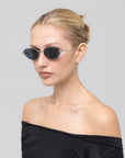 A woman with blonde hair in a bun wears Rebirth sunglasses by For Art's Sake®, paired with an off-the-shoulder black top, posing against a plain light gray background.