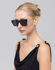 A blonde woman wears For Art's Sake® Incomplete black square acetate sunglasses with gold arms, paired with a black sleeveless top with gold chain straps, set against a plain light gray background.
