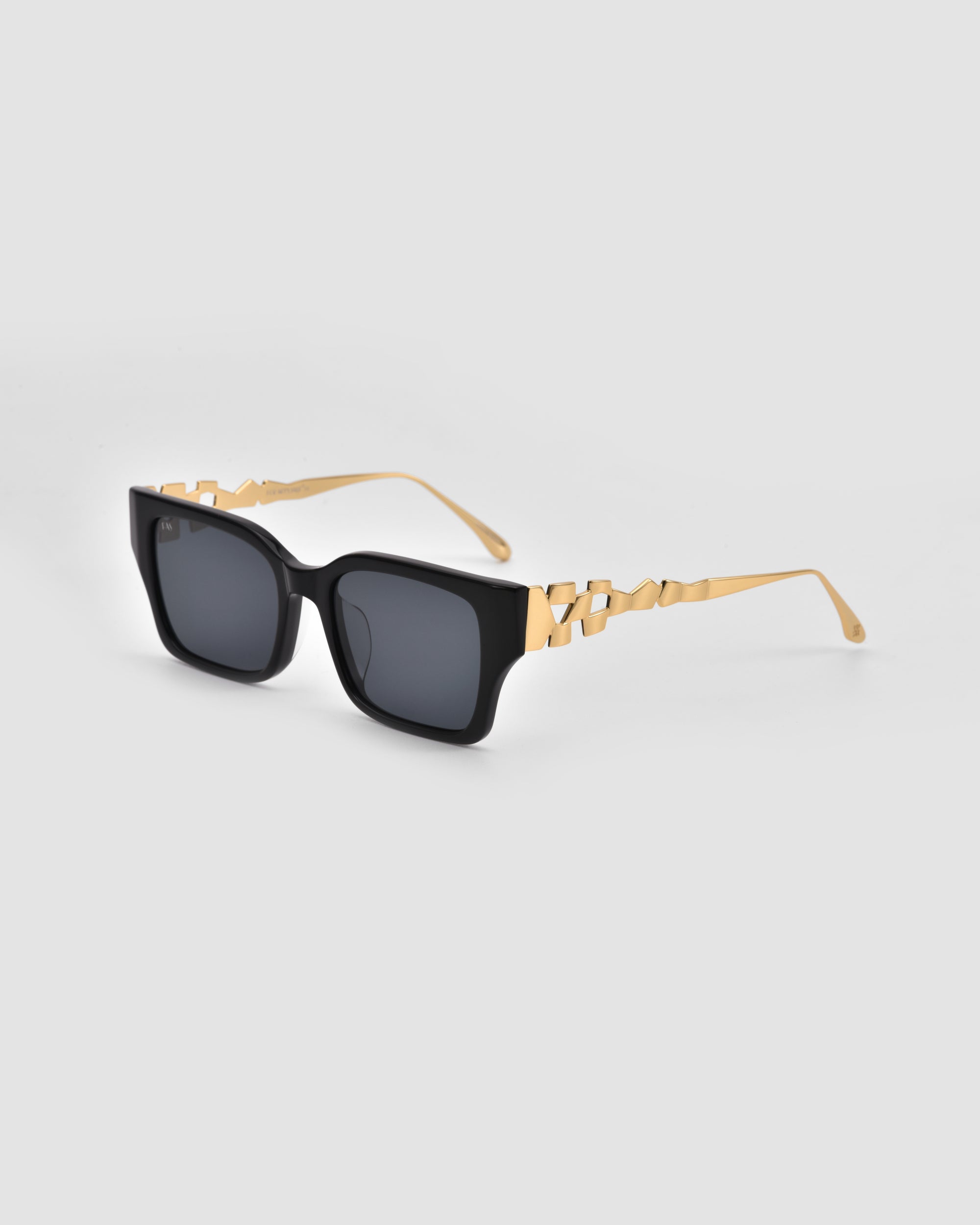 The Incomplete sunglasses by For Art's Sake® feature black boxy frames and gold geometric mosaic metal arms, shown on a light gray background.