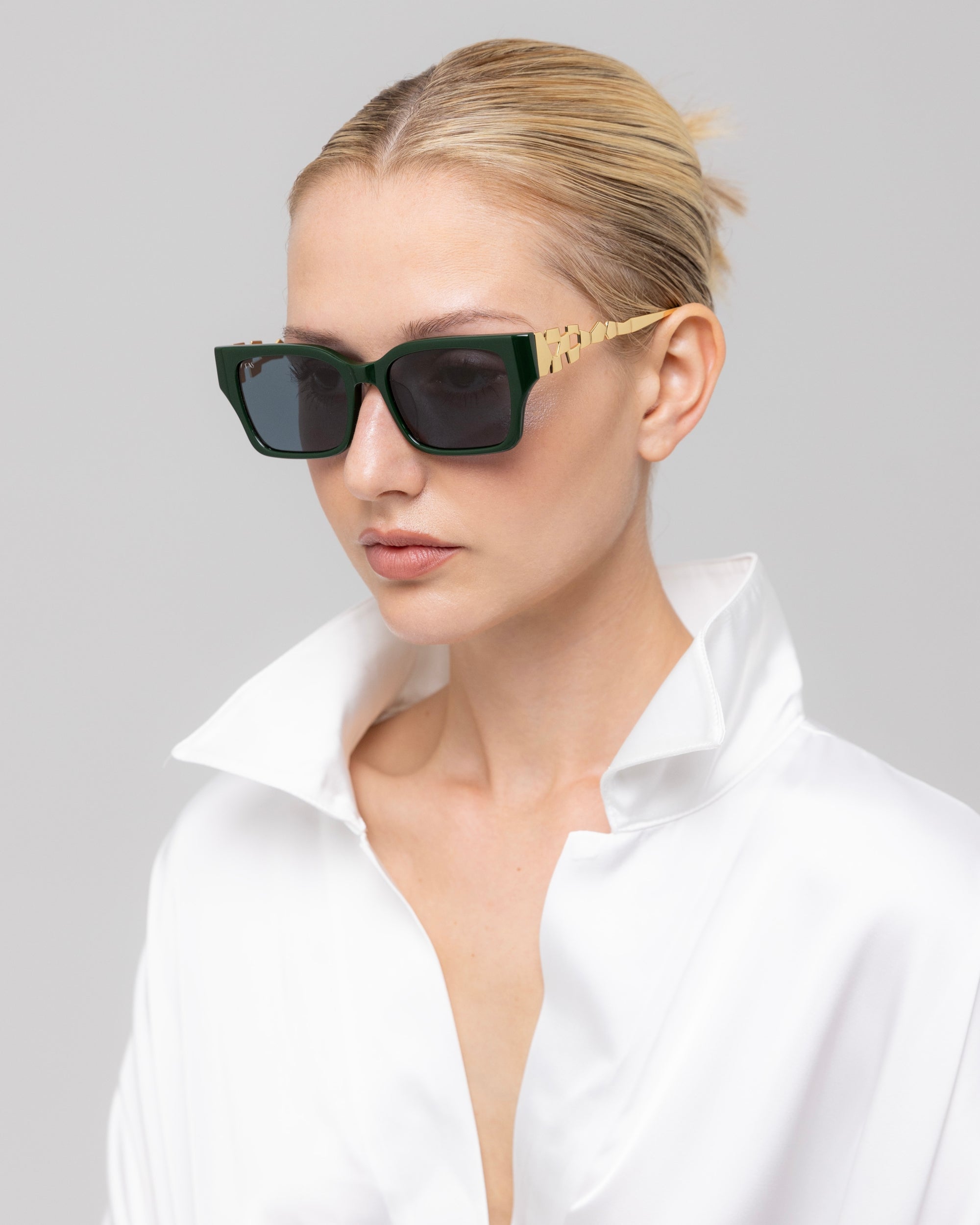 A woman with blonde hair in a neat bun models For Art's Sake® Incomplete sunglasses featuring gold arms and bold, boxy frames, styled with a white satin shirt against a light gray backdrop.