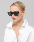 A woman with blonde hair in a neat bun models For Art's Sake® Incomplete sunglasses featuring gold arms and bold, boxy frames, styled with a white satin shirt against a light gray backdrop.