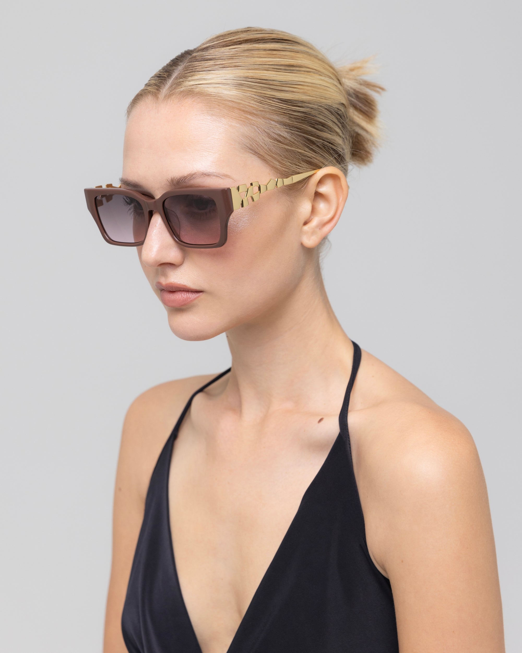 A woman with blonde hair in a neat bun wears For Art's Sake® Incomplete square acetate sunglasses with gold mosaic metal arms and a black halter top, posing against a plain light gray background.