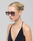 A woman with blonde hair in a neat bun wears For Art's Sake® Incomplete square acetate sunglasses with gold mosaic metal arms and a black halter top, posing against a plain light gray background.