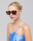 A woman with blonde hair in a bun models the For Art's Sake® Incomplete large square acetate sunglasses and a shiny blue spaghetti strap top with a rosette, posing against a light gray background.