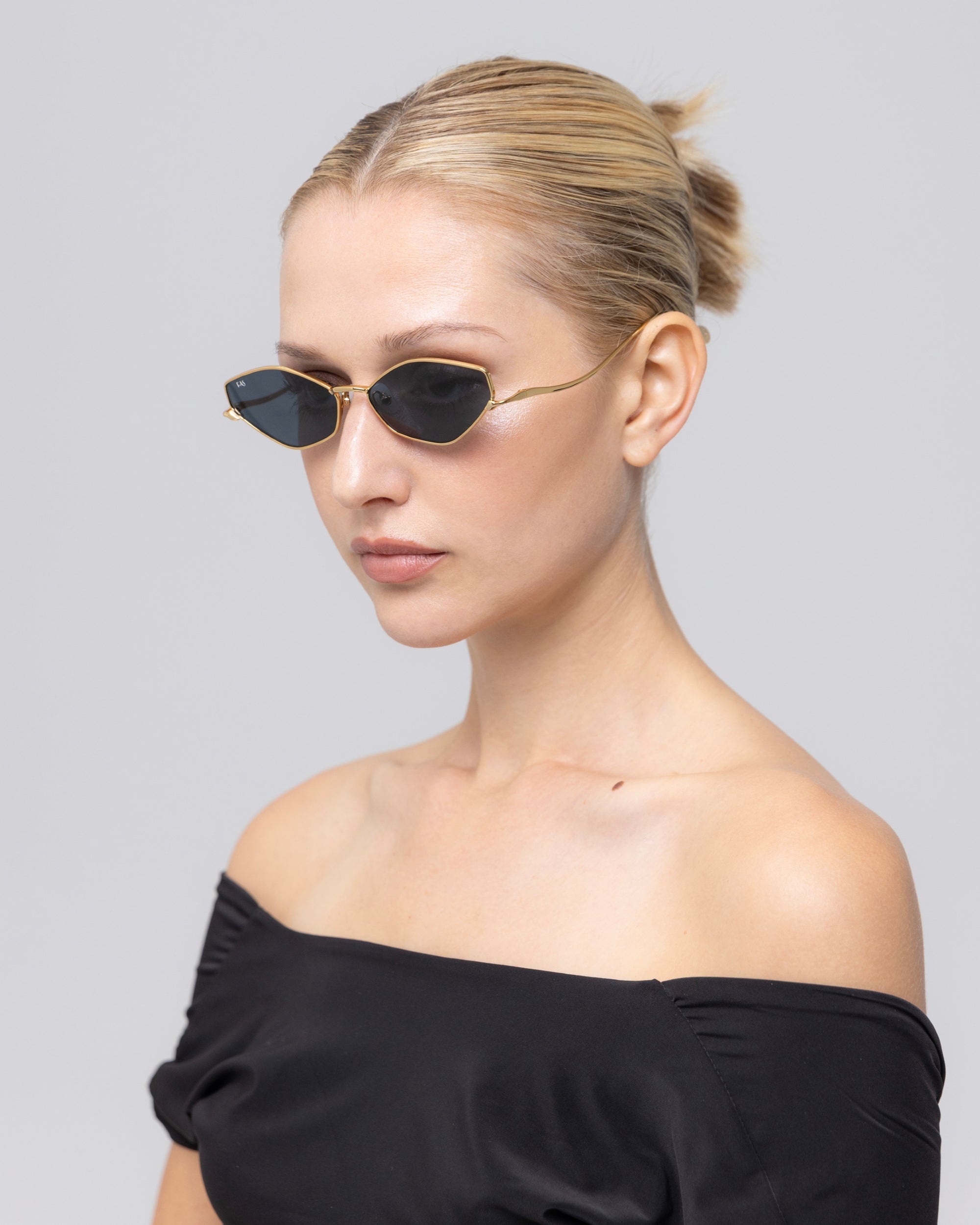 A woman with blonde hair in a sleek bun wears For Art's Sake® Moment small oval black metal sunglasses and a black off-the-shoulder top against a plain light gray background.