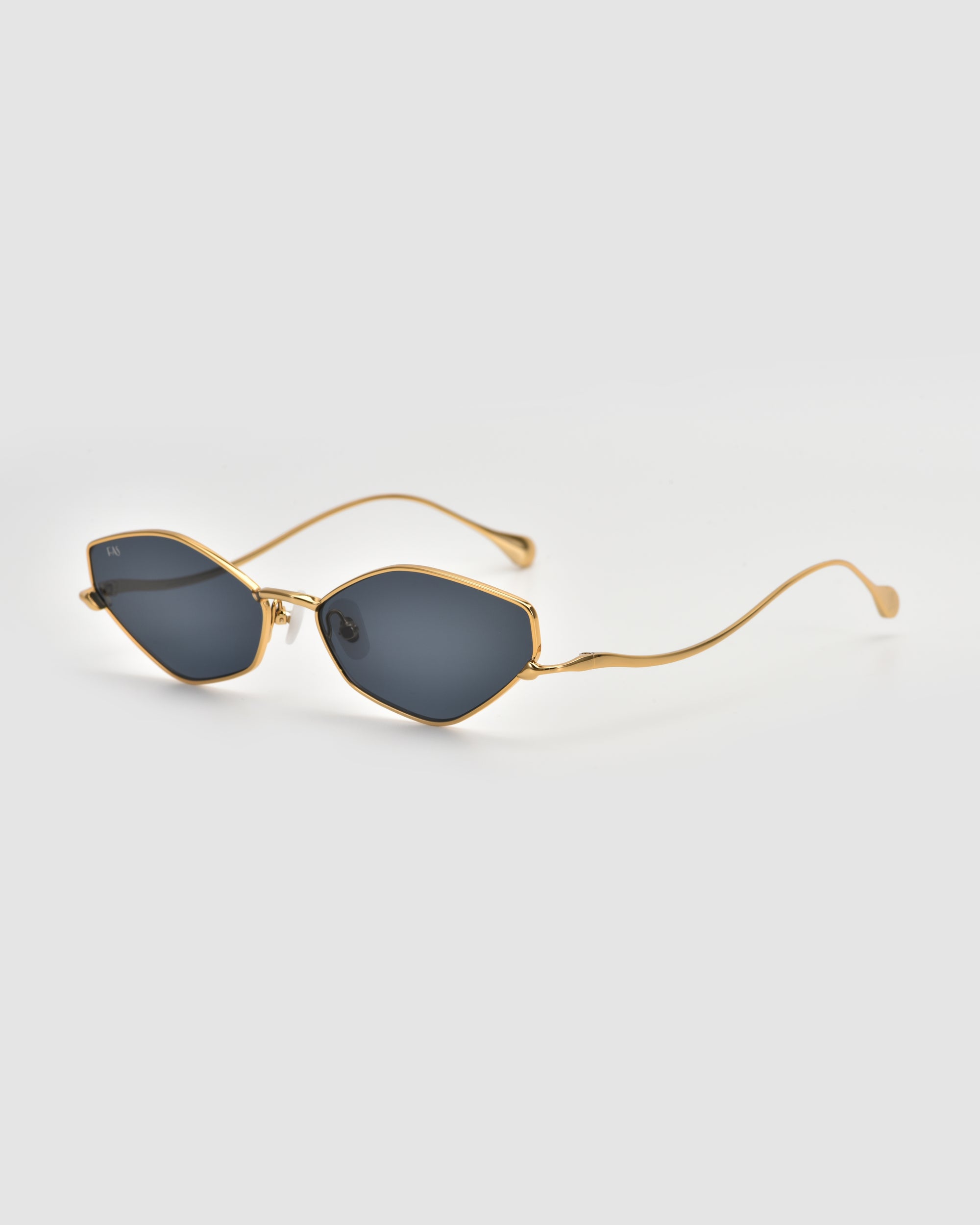 The Moment sunglasses by For Art's Sake® feature a geometric silhouette, dark oval lenses, and a slim gold metal frame with curved arms, displayed on a plain light gray background.