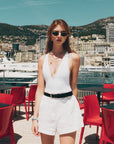 Wearing For Art's Sake® Moment sunglasses, a woman in a white sleeveless top and shorts stands on a sunny terrace with red chairs, yachts, and a city hillside behind her.
