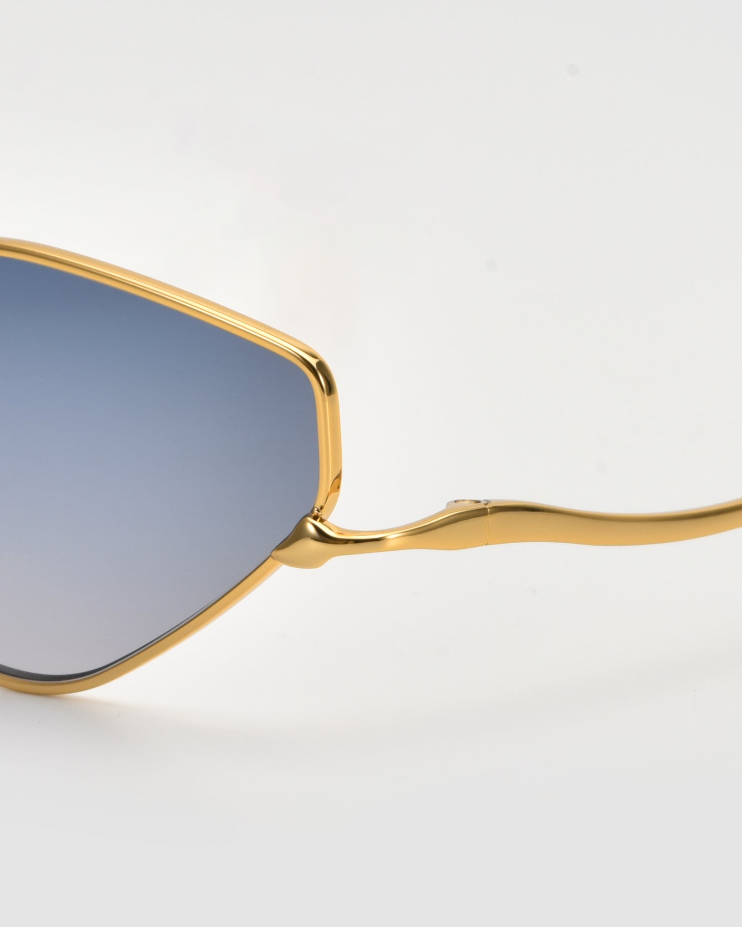 Close-up of For Art&#39;s Sake® Moment sunglasses, showcasing a gold geometric frame and blue-tinted lenses against a light background, with only part of the lens and frame in view.