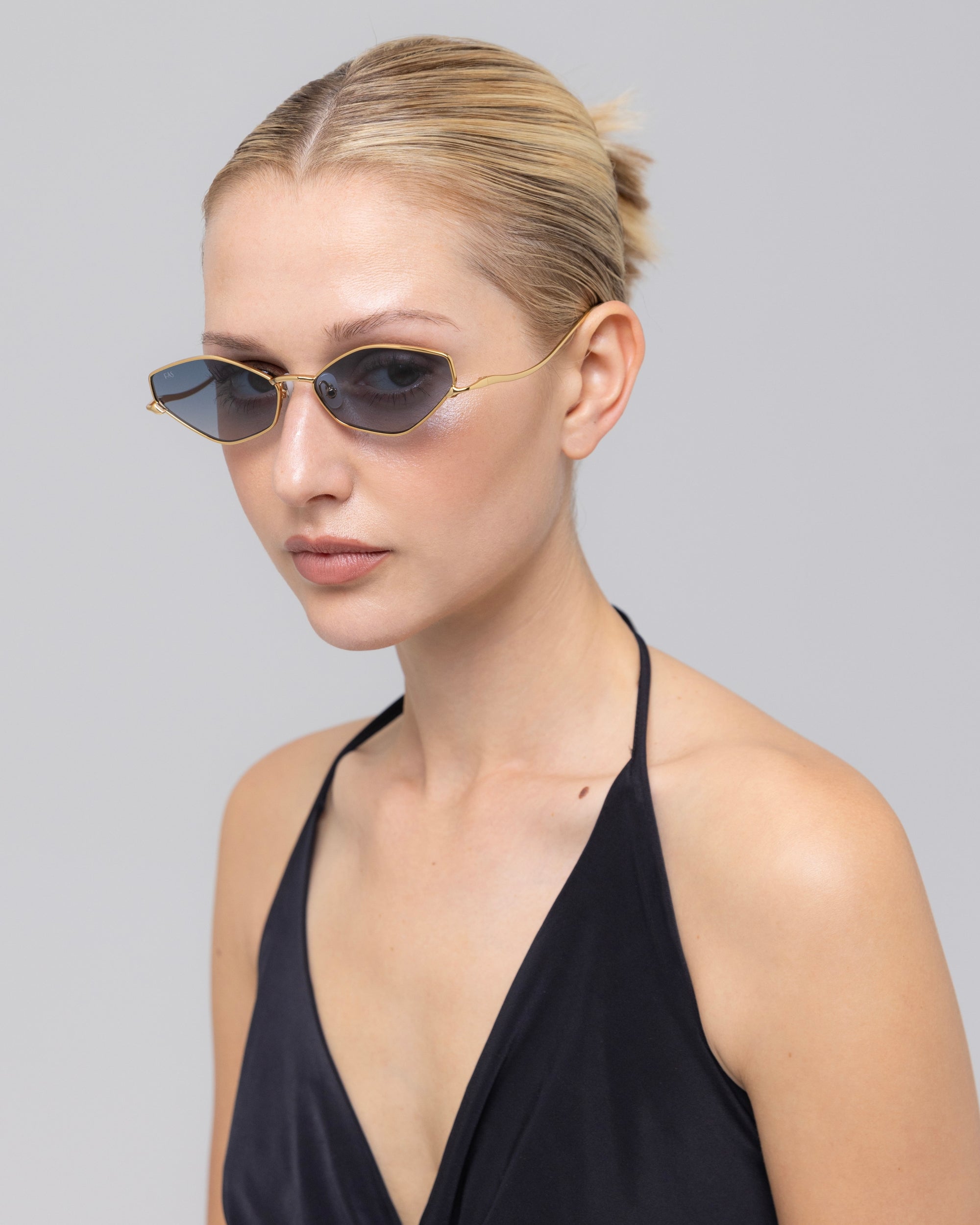 A woman with blonde hair in a neat bun wears For Art’s Sake® Moment sunglasses—small, oval with a sleek metal frame—and a black halter-neck top, looking slightly to the side against a plain light gray background.