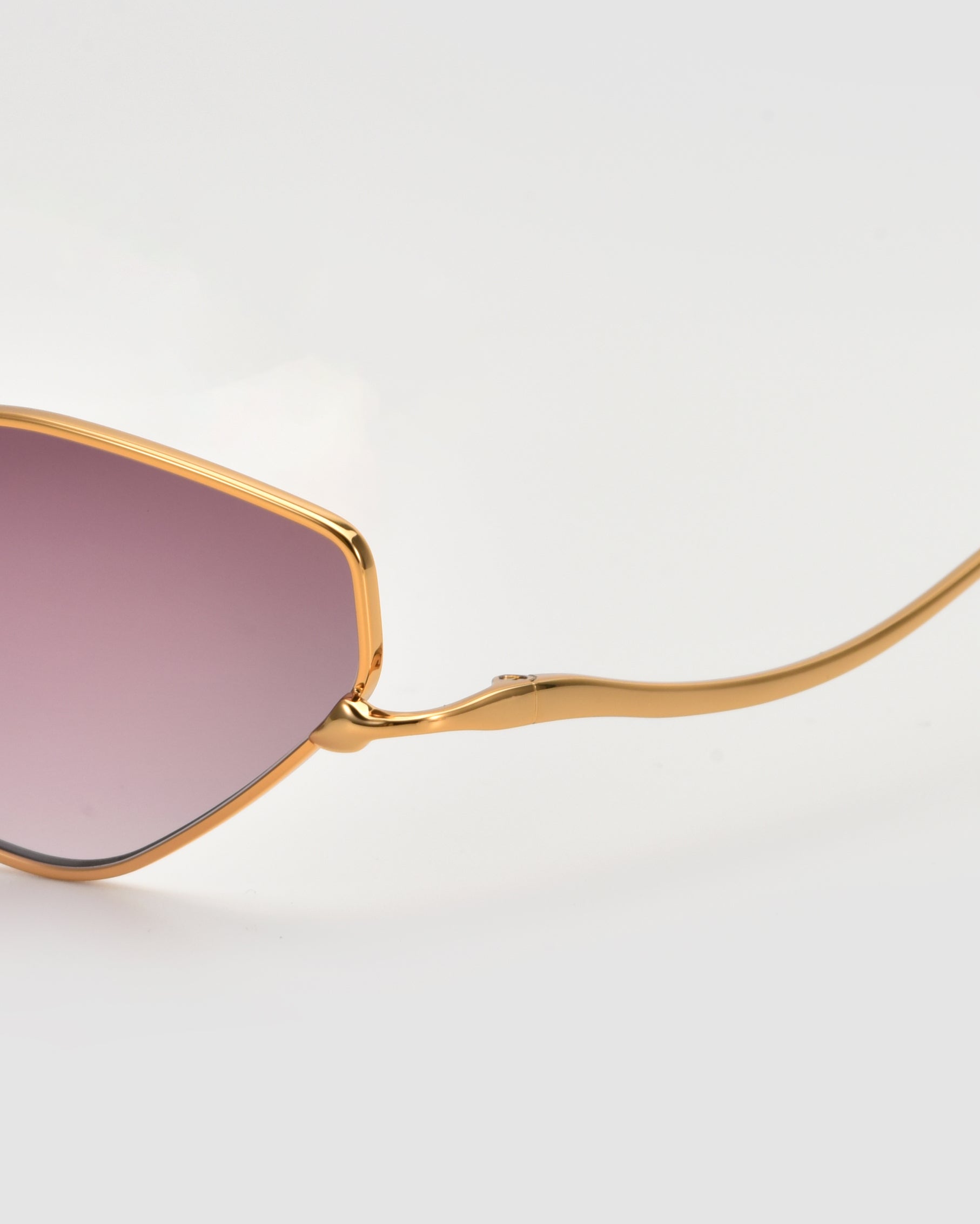 Close-up of the Moment sunglasses by For Art&#39;s Sake® featuring a gold metal frame, purple-tinted lenses, and a sleek geometric arm silhouette against a plain light background.