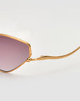 Close-up of the Moment sunglasses by For Art's Sake® featuring a gold metal frame, purple-tinted lenses, and a sleek geometric arm silhouette against a plain light background.