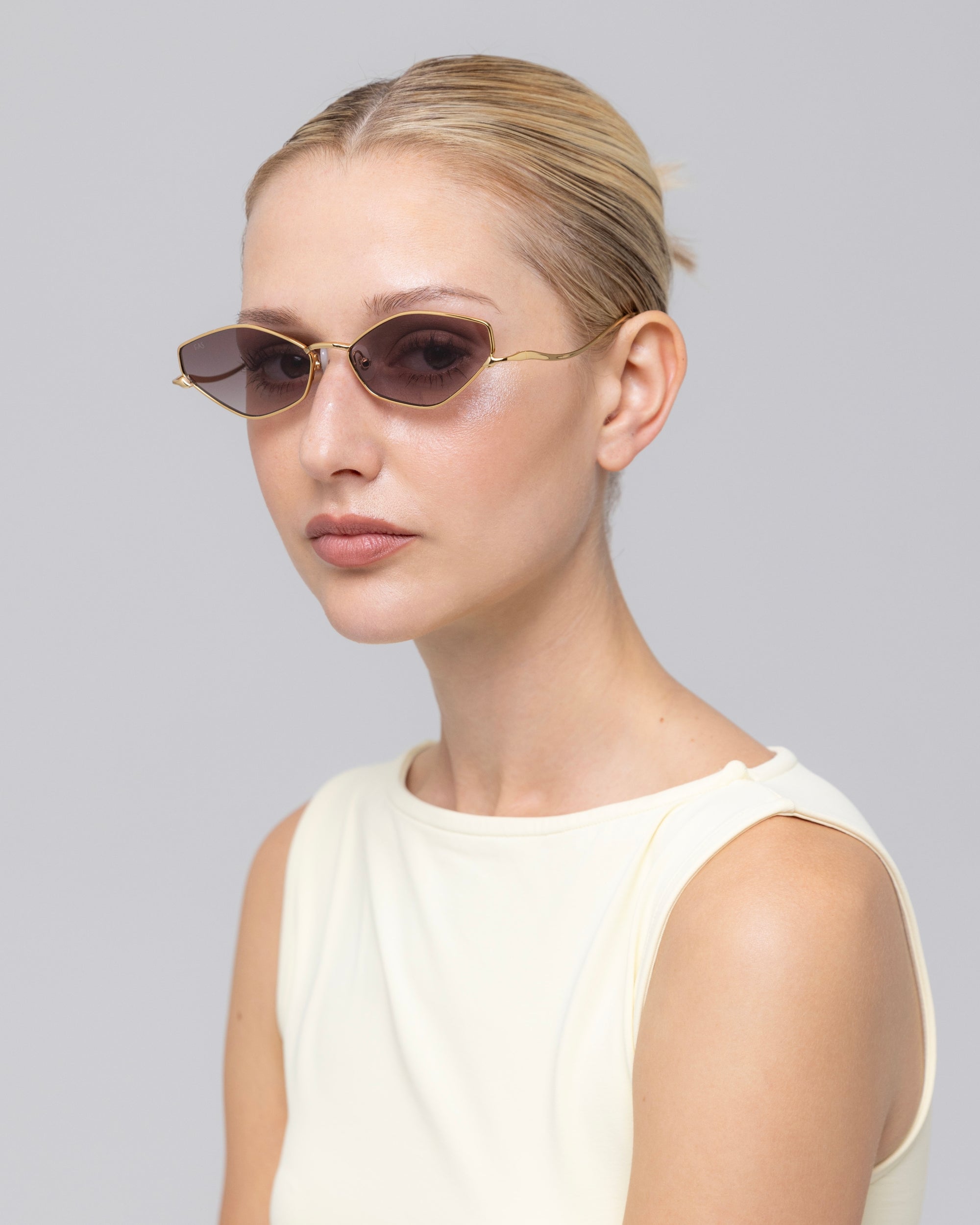 A woman with blonde hair in a low bun wears For Art's Sake® Moment slim, oval metal-frame sunglasses and a light sleeveless top, gazing to the side against a neutral background.