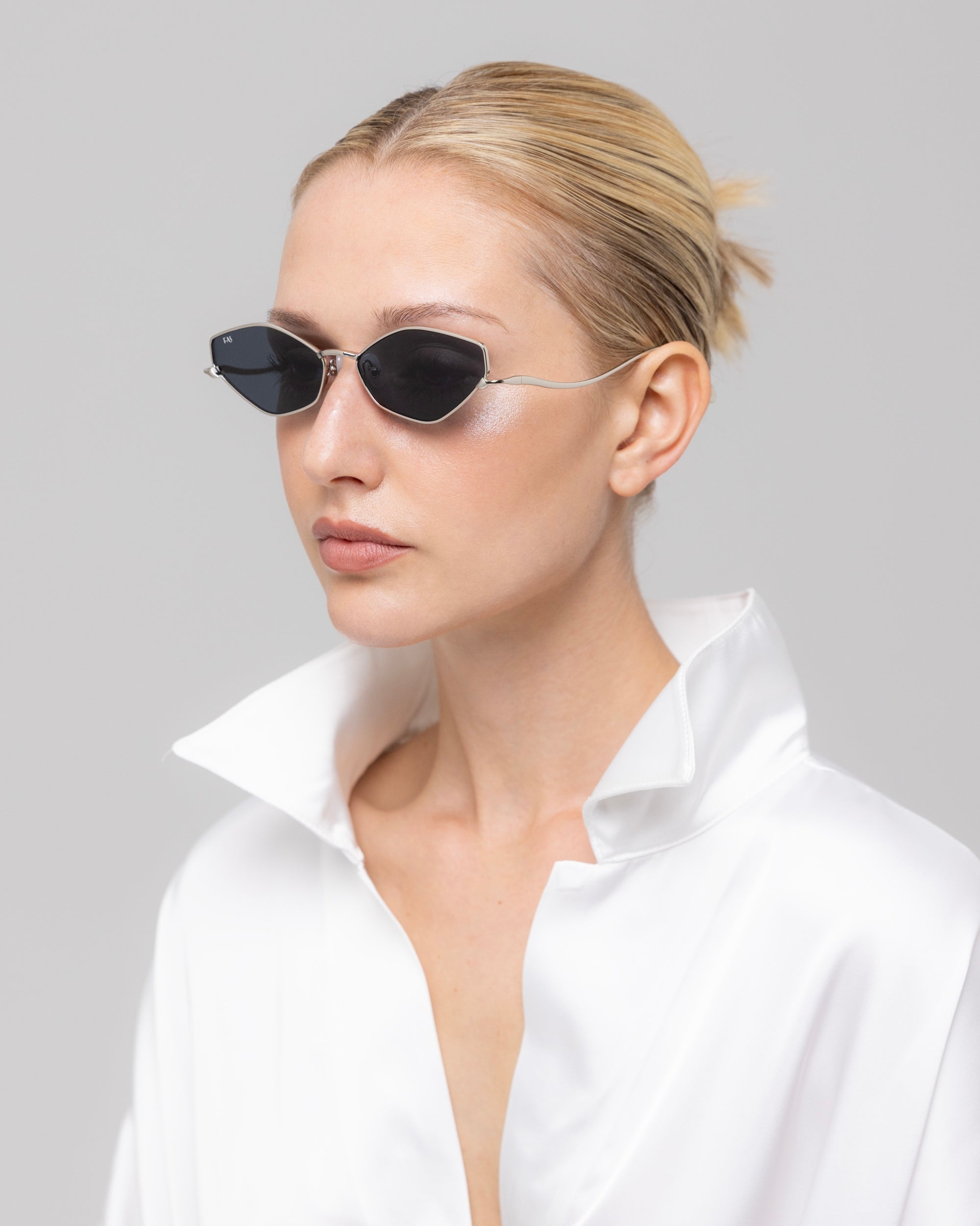 A woman with blonde hair in a sleek bun wears the Moment sunglasses by For Art's Sake®—narrow black metal frames—paired with a shiny white high-collar shirt, creating a geometric silhouette against a light gray background.
