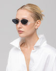 A woman with blonde hair in a sleek bun wears the Moment sunglasses by For Art's Sake®—narrow black metal frames—paired with a shiny white high-collar shirt, creating a geometric silhouette against a light gray background.