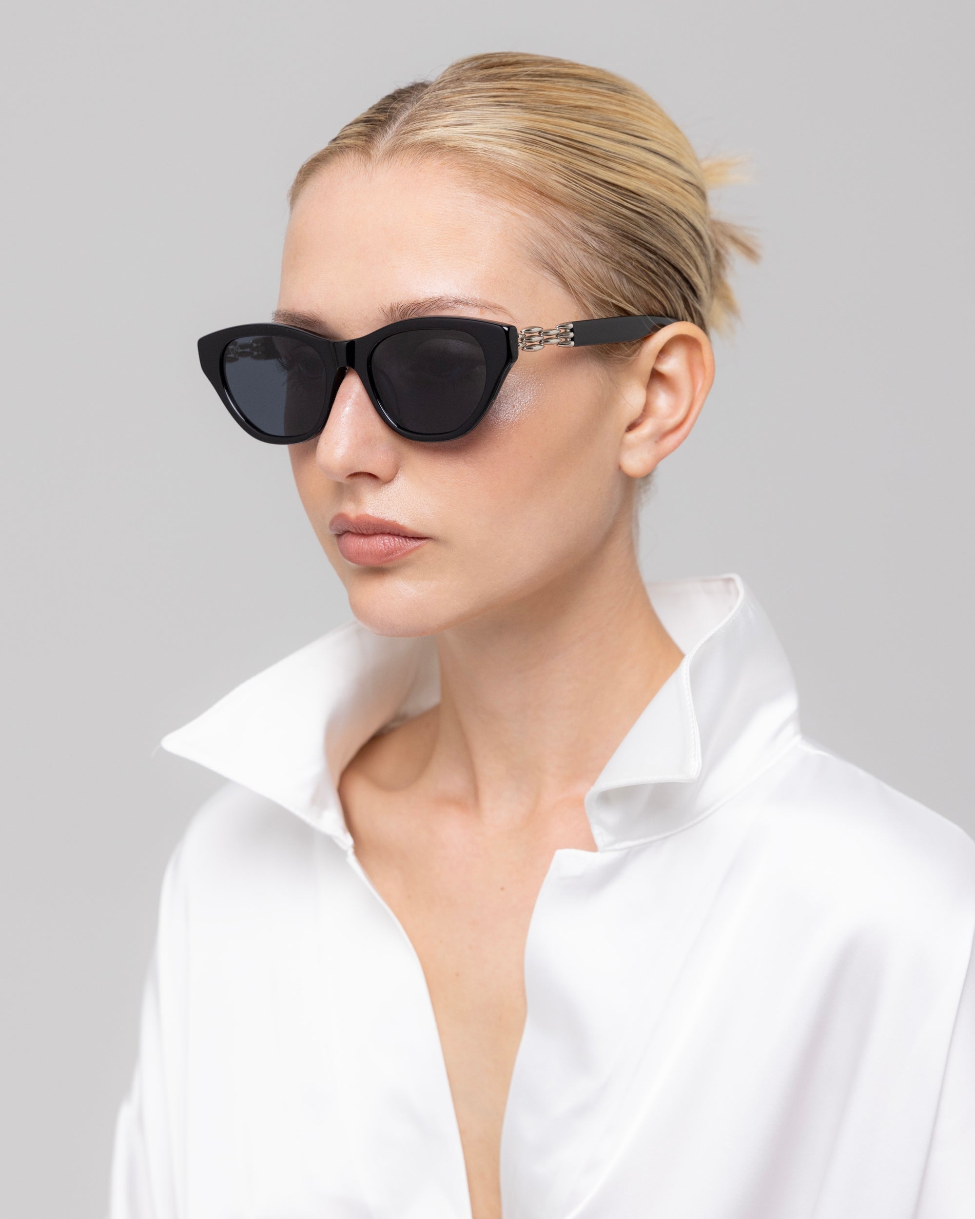 A woman with slicked-back blonde hair models The Cuts sculptural cat-eye sunglasses by For Art's Sake®, paired with a white satin shirt, gazing off to the side against a plain light background.