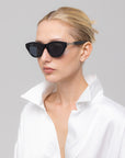 A woman with slicked-back blonde hair models The Cuts sculptural cat-eye sunglasses by For Art's Sake®, paired with a white satin shirt, gazing off to the side against a plain light background.