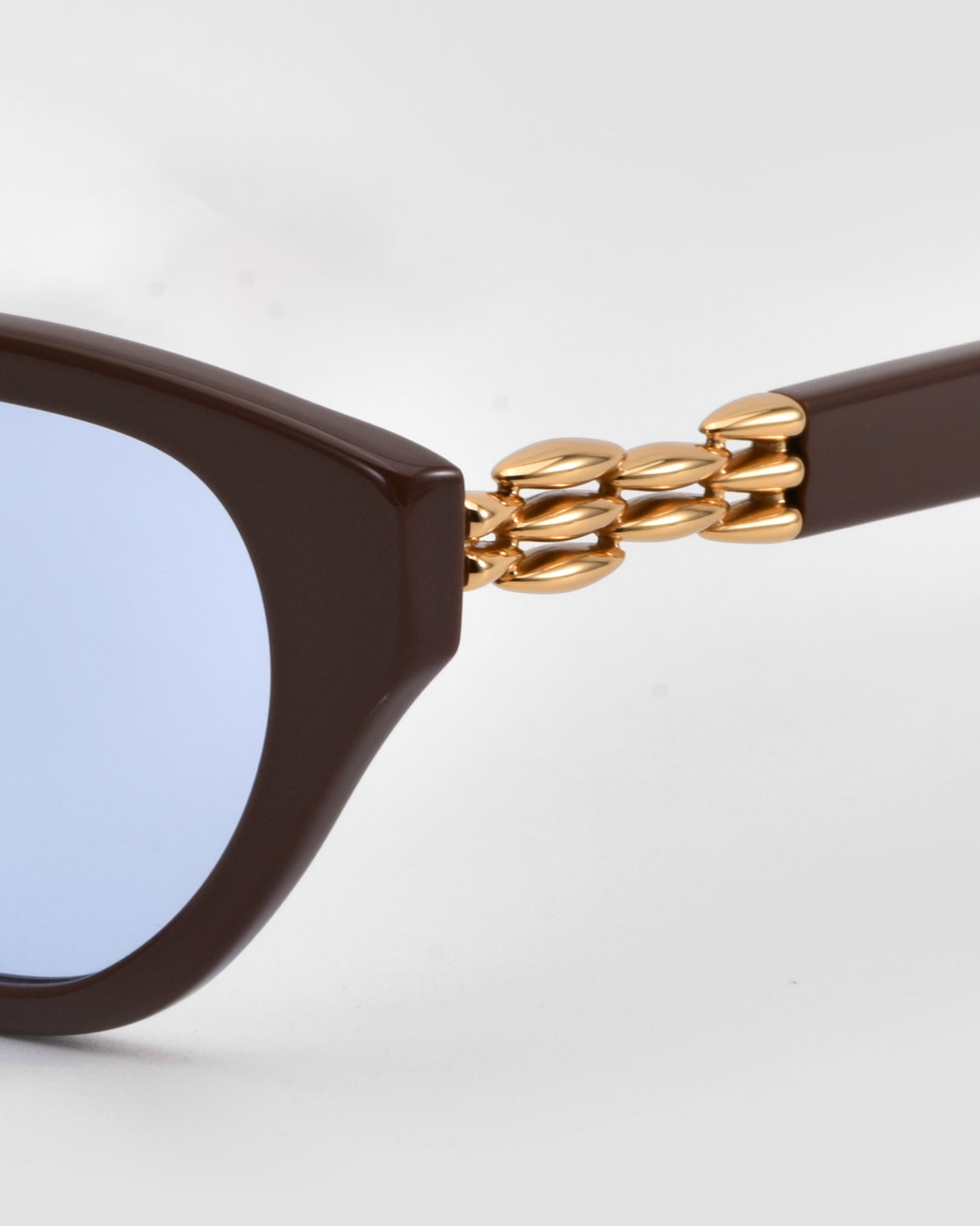 Close-up of The Cuts by For Art&#39;s Sake®—sculptural sunglasses with a weaved metal frame, brown arms, blue-tinted lenses, and gold chain-like temple details, set against a light gray background.