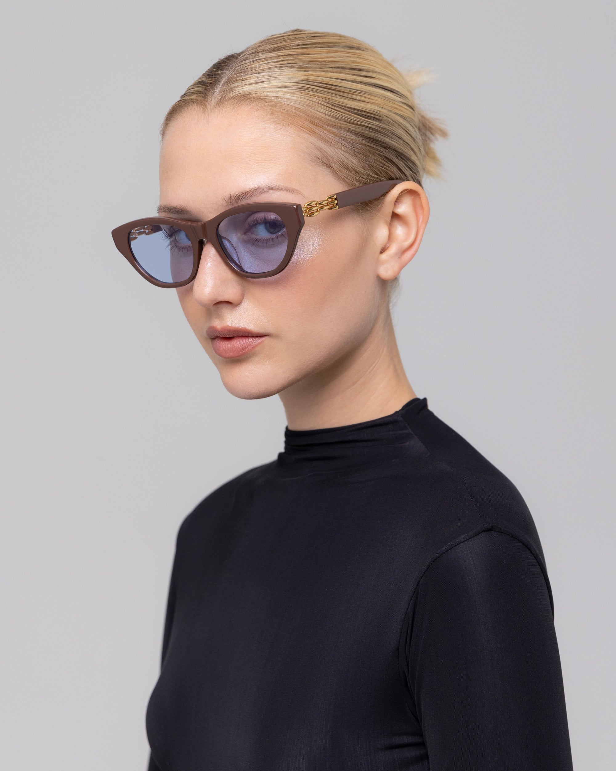 A blonde woman with a neat bun wears The Cuts cat-eye sunglasses by For Art's Sake® and a black high-neck top, posing against a plain gray background.