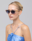 A woman with blonde hair in a neat bun models The Cuts sunglasses by For Art's Sake®, paired with a shiny, light blue spaghetti strap top featuring a rose detail, set against a plain grey background.
