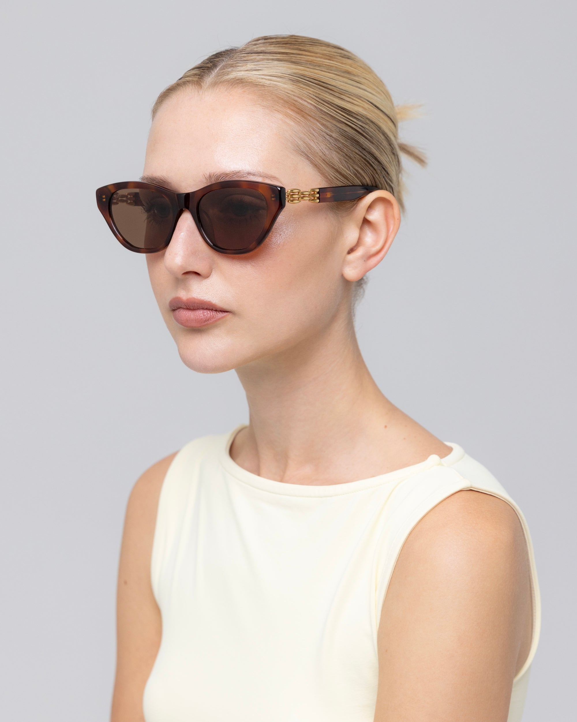 A woman with blonde hair pulled back models The Cuts cat-eye sunglasses by For Art's Sake®, paired with a sleeveless cream top against a plain light gray background.
