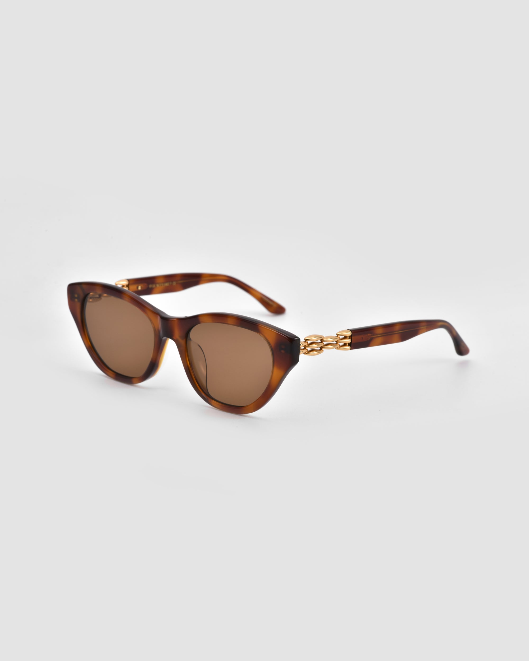 The Cuts sunglasses by For Art's Sake® feature a tortoiseshell sculptural frame, round brown lenses, and gold chain accents on the arms, shown against a plain light gray background.