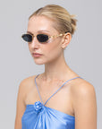 A woman with blonde hair in a sleek bun wears For Art's Sake® Droplets retro oval sunglasses with gold frames and a shiny blue satin top, posing against a plain light gray background.