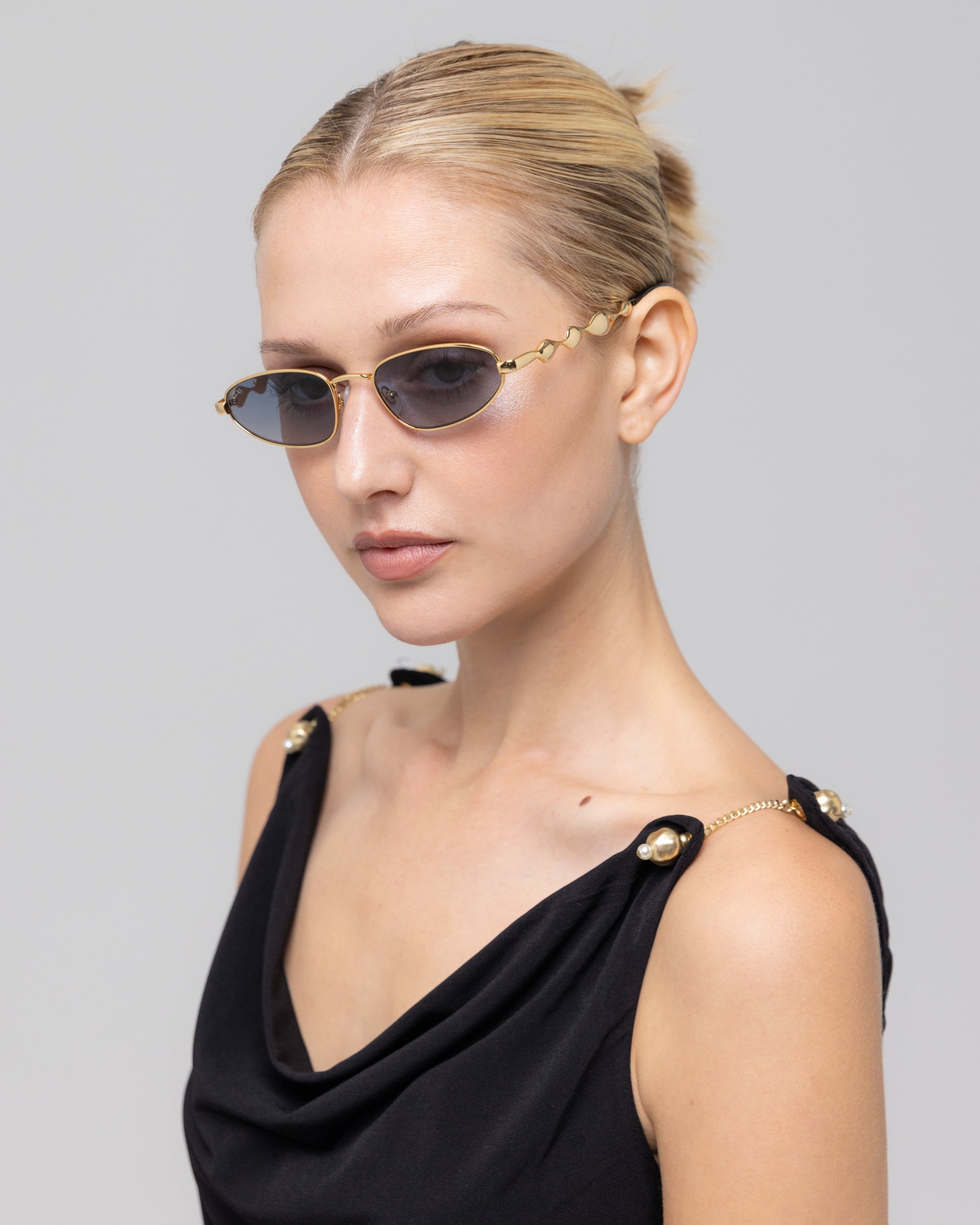 A woman with blonde hair in a sleek bun wears For Art's Sake® Droplets retro oval sunglasses and a black sleeveless top with gold chain and button details, posing against a light gray background.