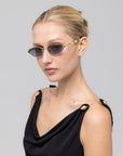 A woman with blonde hair in a sleek bun wears For Art's Sake® Droplets retro oval sunglasses and a black sleeveless top with gold chain and button details, posing against a light gray background.