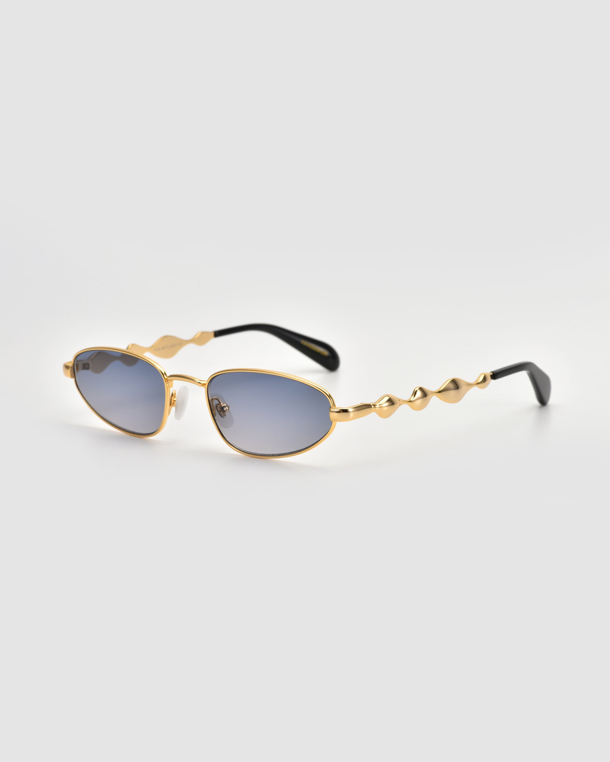 Droplets by For Art's Sake®: gold metal frame sunglasses with wavy sculpted arms and oval blue-tinted lenses, shown on a light gray background for a subtle retro vibe.
