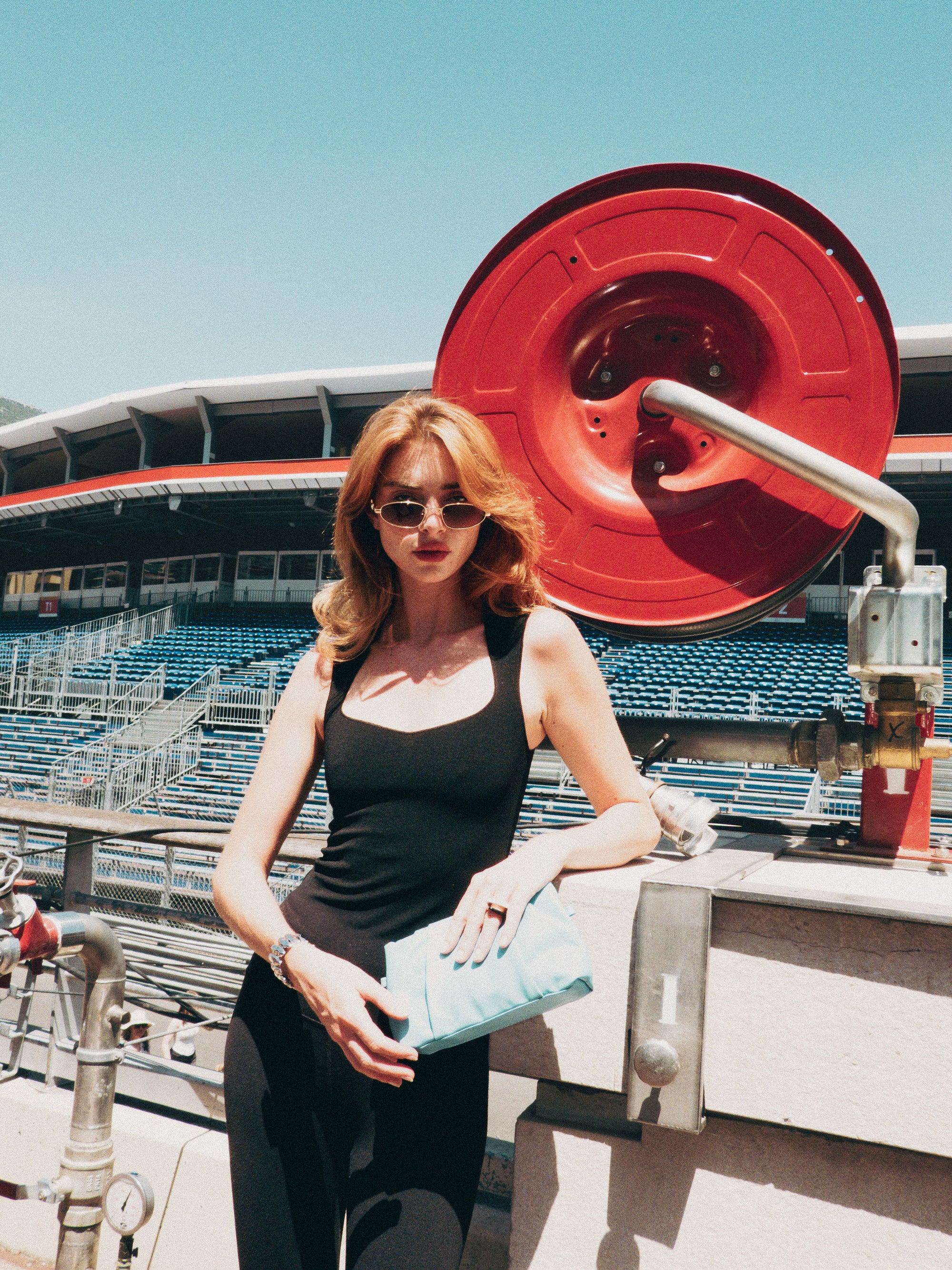 A woman in a black tank top and For Art's Sake® Droplets retro sunglasses stands by a red industrial reel at an outdoor stadium, holding a light blue clutch with empty bleachers behind her.