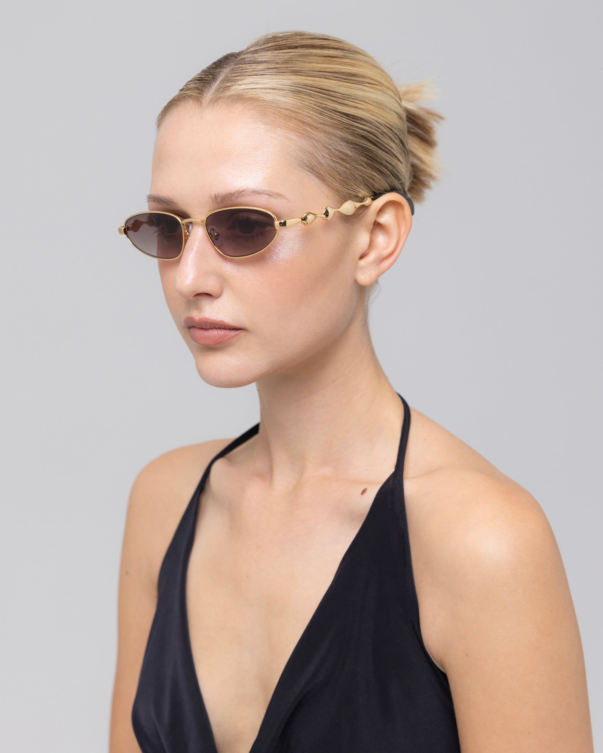 A woman with blond hair in a sleek bun wears For Art's Sake® Droplets sunglasses—narrow, retro oval gold frames—paired with a black halter top, posing against a plain light gray background.