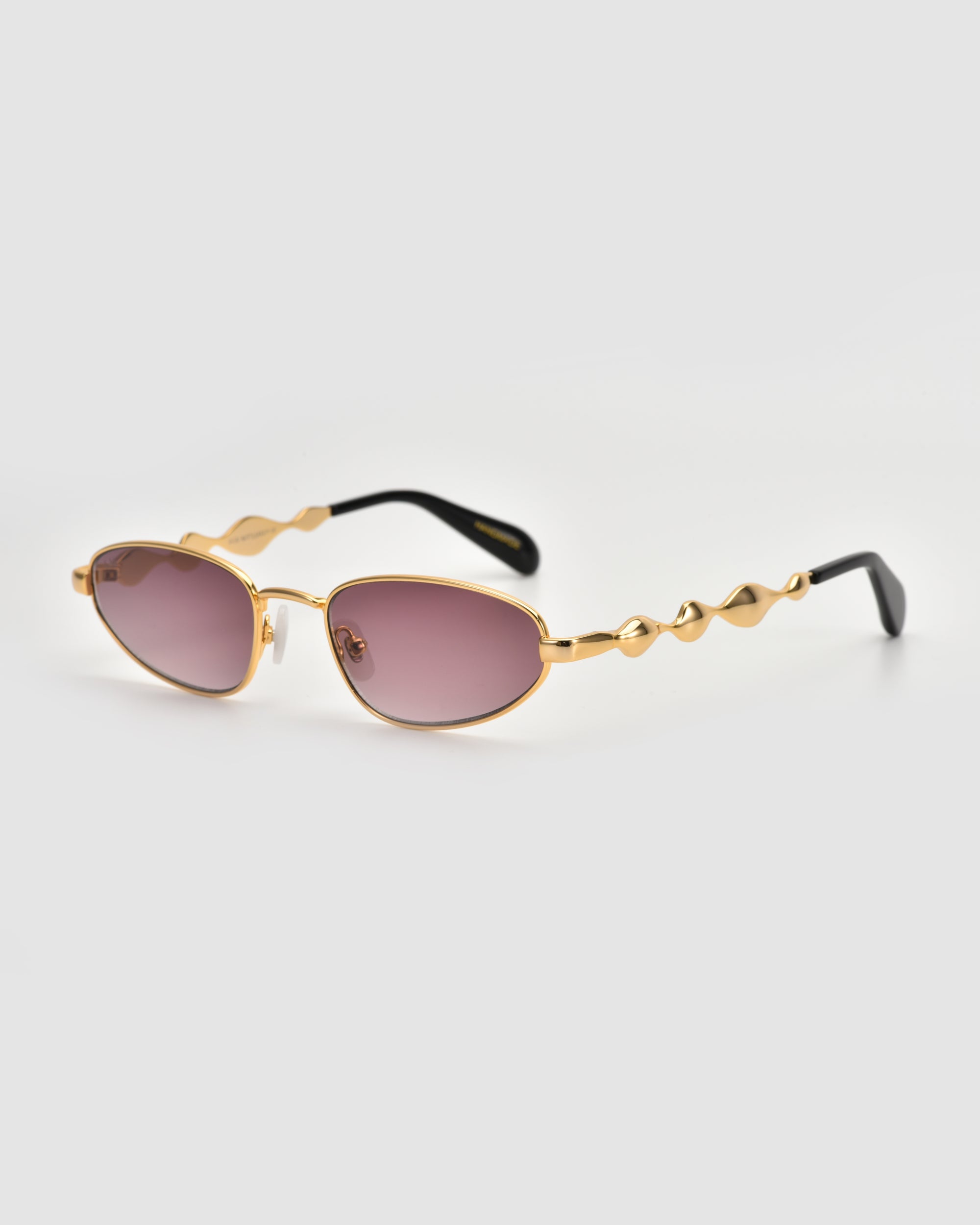 Droplets by For Art's Sake®: Gold-framed, retro oval sunglasses with rose-tinted lenses, wavy sculpted metal temples, and sleek black earpieces. Shown on a plain light gray background.