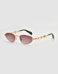 Droplets by For Art's Sake®: Gold-framed, retro oval sunglasses with rose-tinted lenses, wavy sculpted metal temples, and sleek black earpieces. Shown on a plain light gray background.
