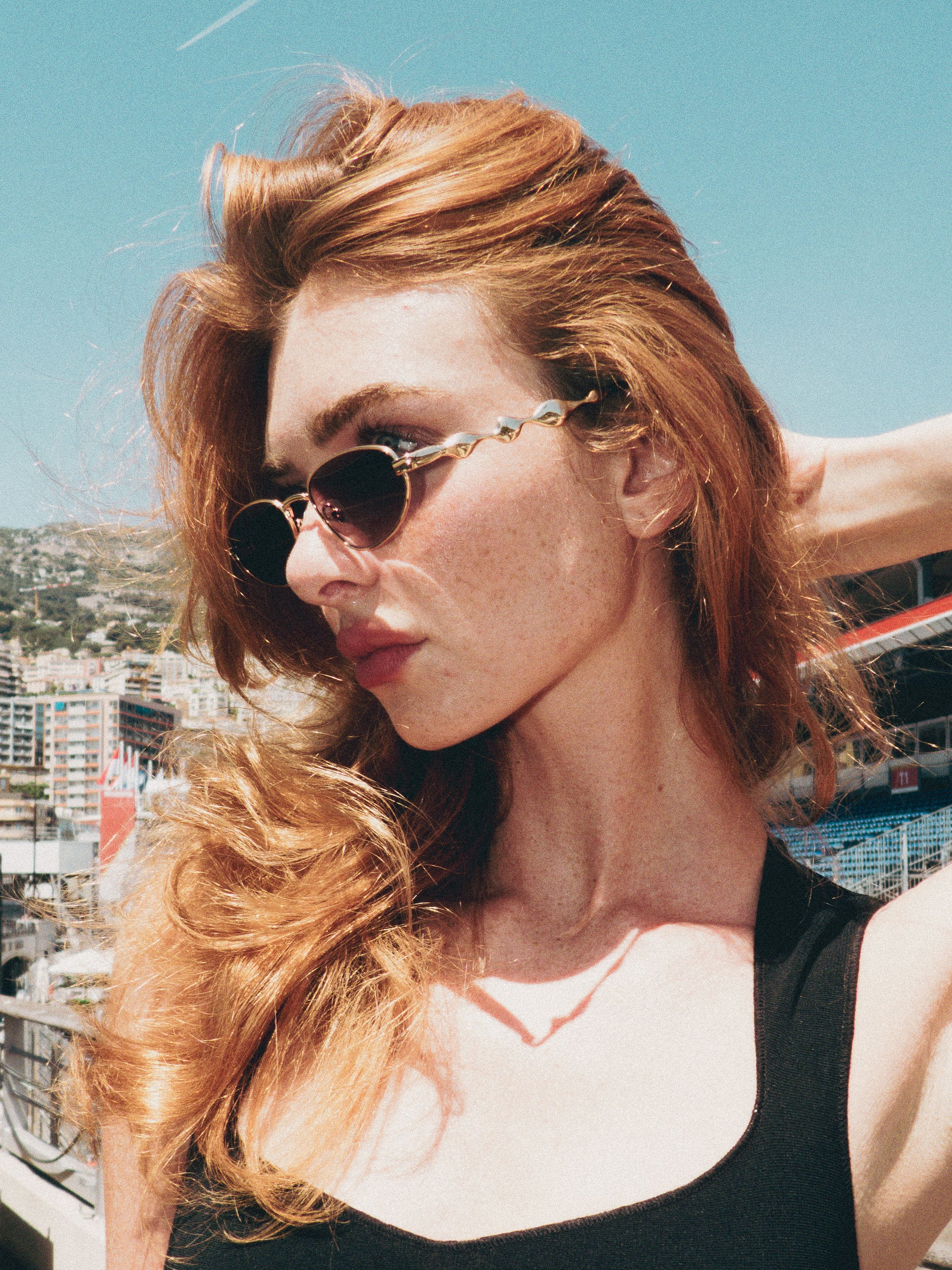 A woman with long red hair models For Art's Sake® Droplets sunglasses and a black top outdoors, posing against a scenic city backdrop under a clear blue sky.