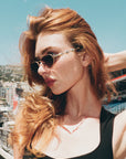 A woman with long red hair models For Art's Sake® Droplets sunglasses and a black top outdoors, posing against a scenic city backdrop under a clear blue sky.