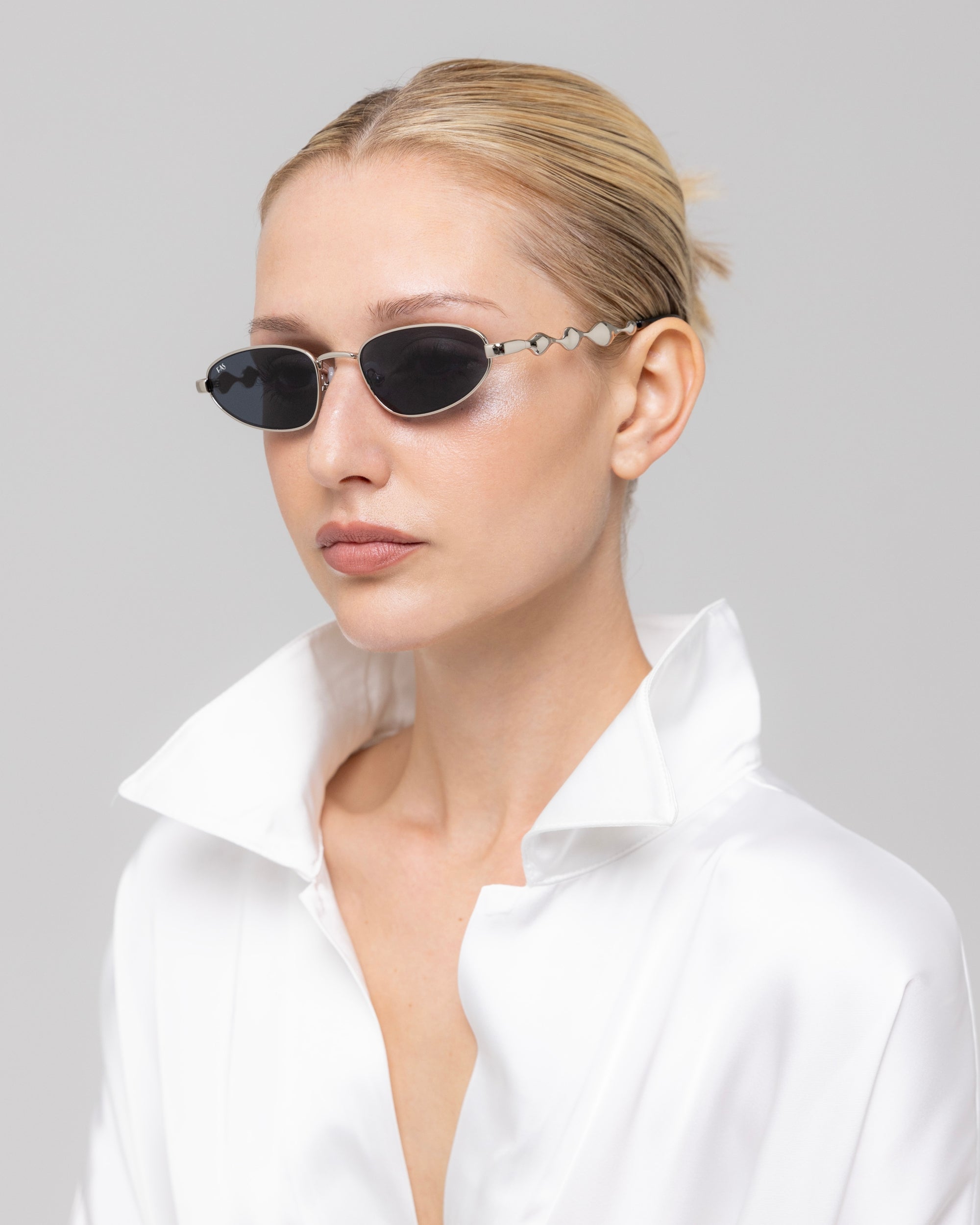 A woman with blonde hair in a slicked-back bun wears For Art's Sake® Droplets narrow retro sunglasses and a white, open-collared shirt, facing slightly sideways against a plain light gray background.