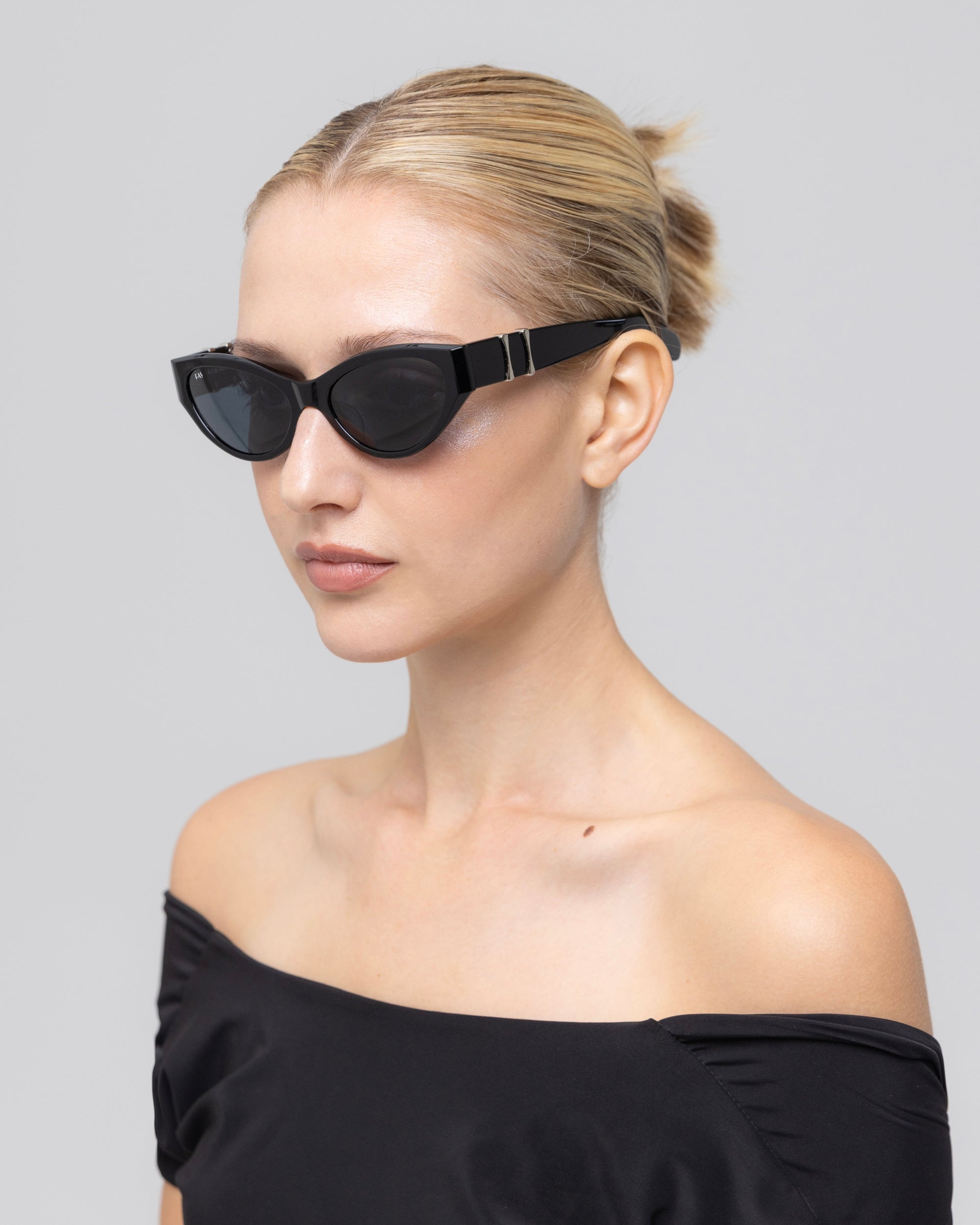 A woman with blonde hair in a sleek bun wears For Art's Sake® Halo(S) black sculptural acetate cat-eye sunglasses and an off-the-shoulder black top, posing against a plain light gray background.