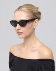 A woman with blonde hair in a sleek bun wears For Art's Sake® Halo(S) black sculptural acetate cat-eye sunglasses and an off-the-shoulder black top, posing against a plain light gray background.