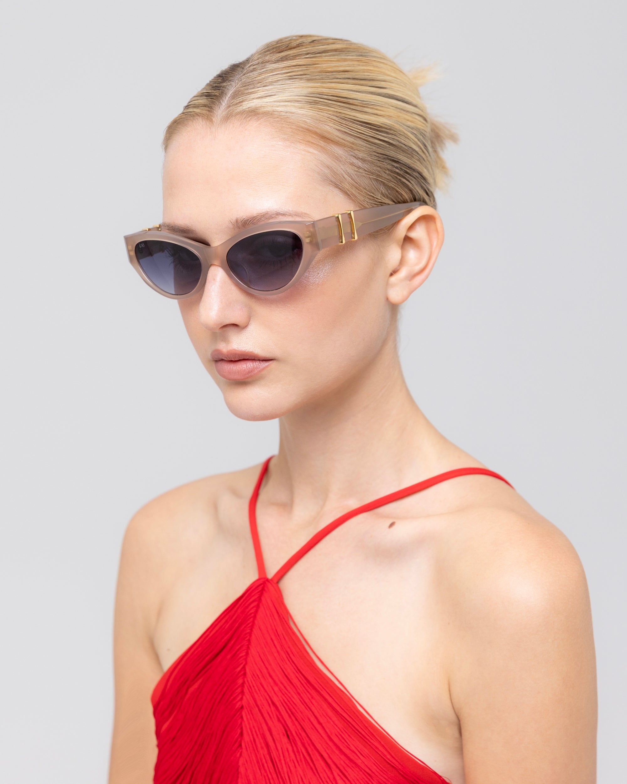 A blonde woman with a low bun wears For Art's Sake® Halo(S) cat-eye sunglasses with a sculptural acetate frame and a red pleated halter-neck top, posed against a plain light gray background.