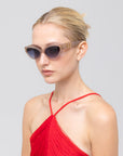 A blonde woman with a low bun wears For Art's Sake® Halo(S) cat-eye sunglasses with a sculptural acetate frame and a red pleated halter-neck top, posed against a plain light gray background.