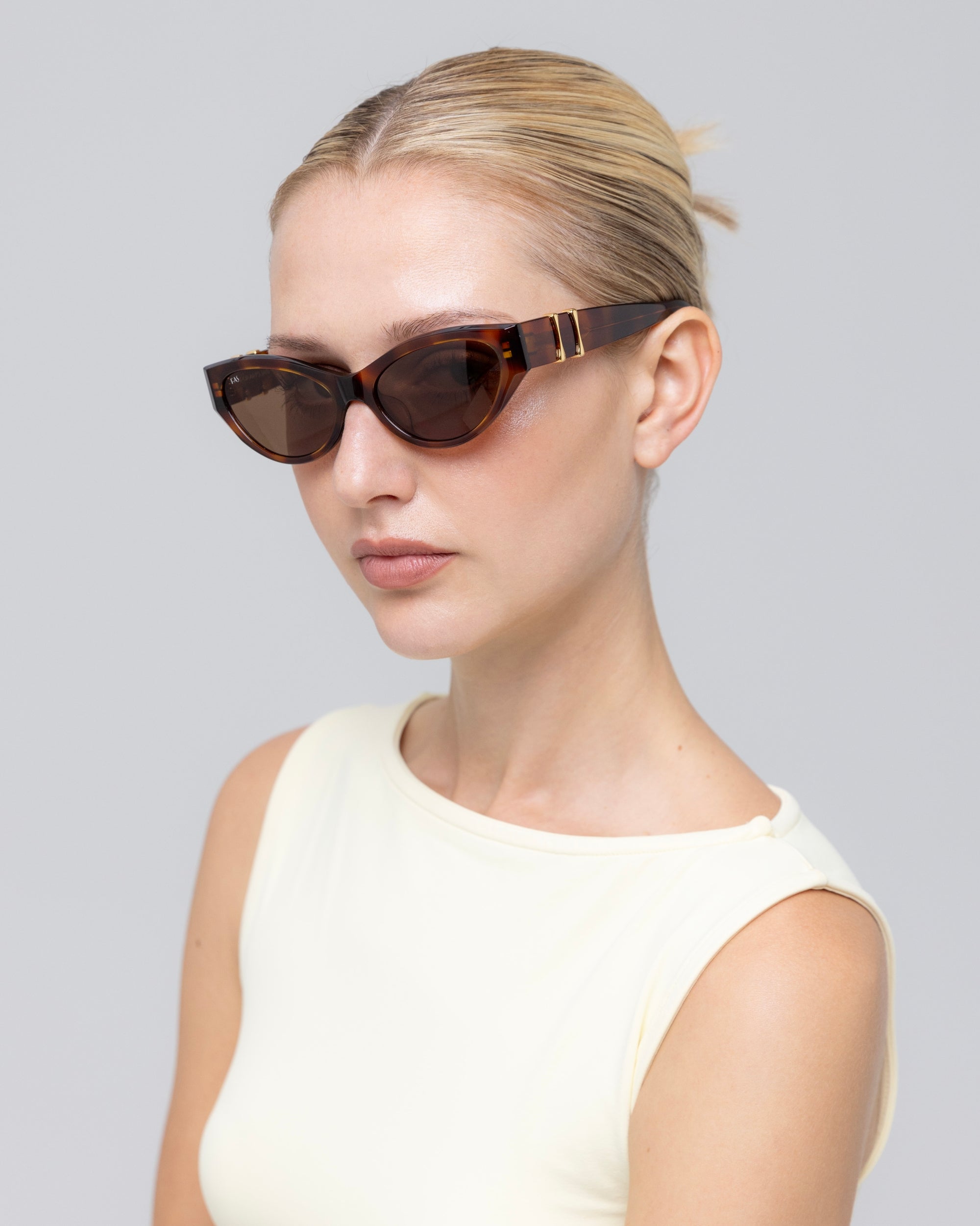 A woman with blonde hair pulled back models For Art's Sake® Halo(S) dark cat-eye sunglasses with a sculptural acetate frame and a sleeveless white top, posing against a plain light gray background.