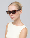 A woman with blonde hair pulled back models For Art's Sake® Halo(S) dark cat-eye sunglasses with a sculptural acetate frame and a sleeveless white top, posing against a plain light gray background.