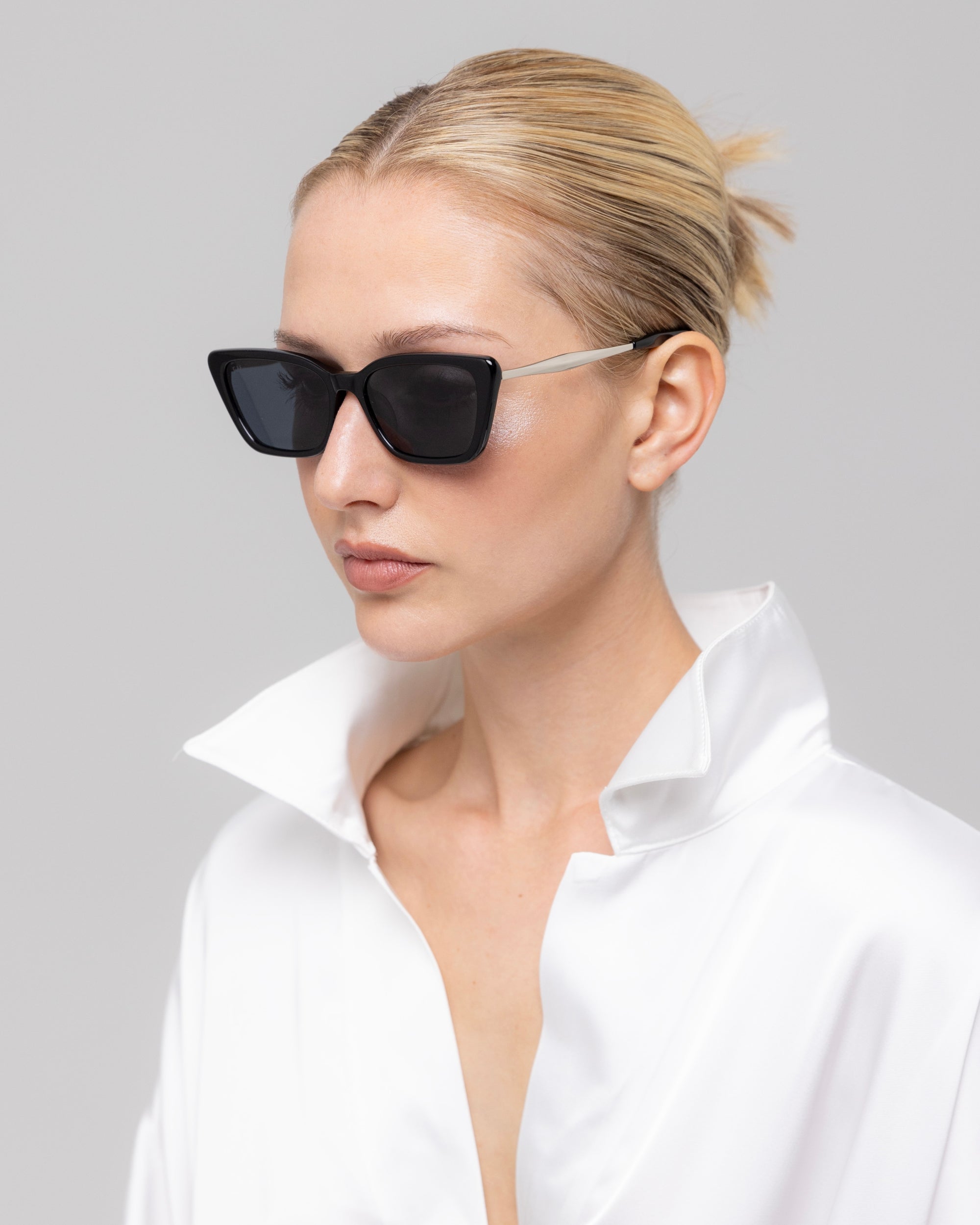 A woman with blonde hair pulled back wears For Art's Sake® Harmony(S) black minimal sunglasses and a white satin shirt with a popped collar, posing against a plain light gray background.